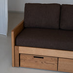 Brown sofa with wooden frame and drawers against a plain wall.